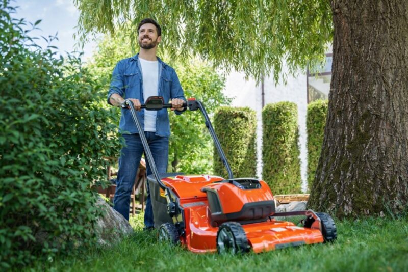 Muškarac kosi travu u dvorištu koristeći motorni trimer kosilicu pored velikog drveta