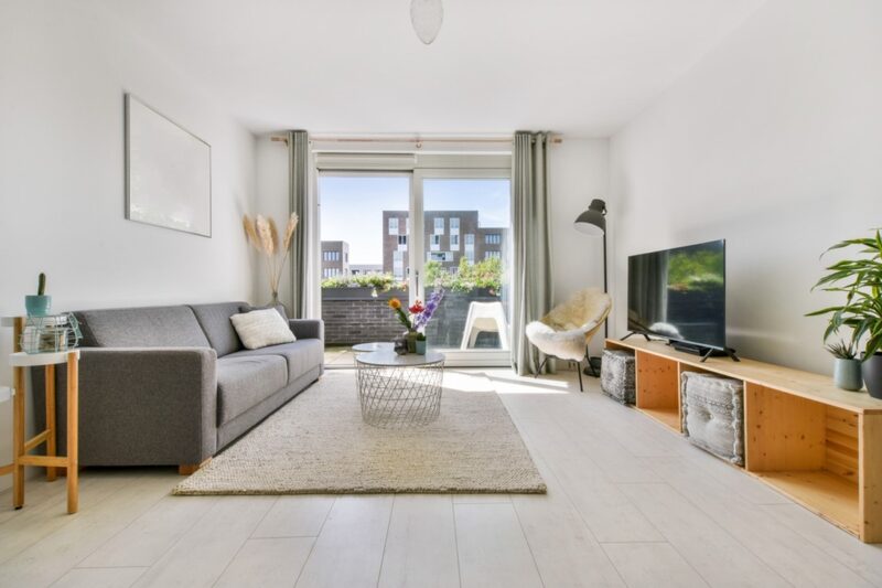 Svetao dnevni boravak sa sivim kaučem, drvenim TV ormarićem, malim stočićem i velikim staklenim vratima koja vode na balkon sa cvećem