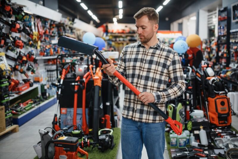 Muškarac u radnji sa alatima drži i pregleda ručni baštenski alat među policama sa opremom