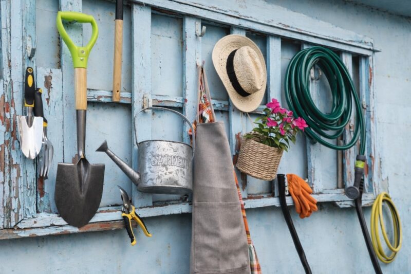 Baštenski alat okačen na starom plavom drvenom zidu, uključujući lopatu, kanticu za zalivanje, šešir, rukavice i crevo