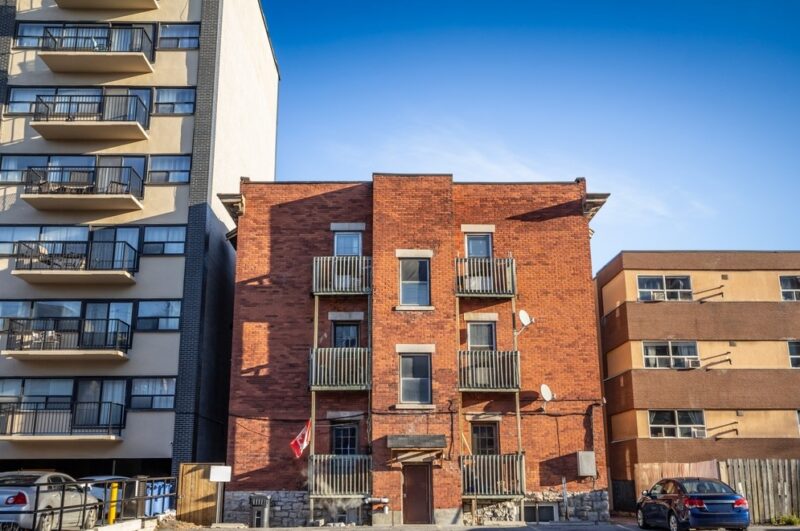 Troetažna zgrada od crvene cigle sa balkonima, smeštena između višespratnice i niže stambene zgrade, pod vedrim nebom